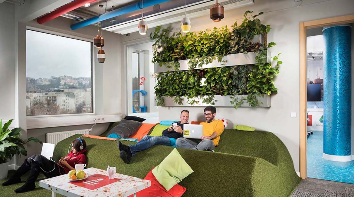 A Google Budapest iroda közösségi tere zöld, füvet imitáló pihenővel, stílusos világítással és növényekkel a falon. Néhány alkalmazott beszélget és relaxál színes párnák között.