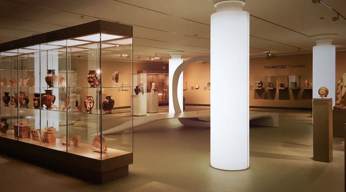 A Szépművészeti Múzeum ókor kiállítása. Antik vázák és edények, valamint más műtárgyak sorakoznak egy impozáns üvegvitrinben az előtérben, még kicsivel hátrébb, fehér, világító oszlopok egy organikus installáció, más vitrinek és szobrok látszanak.