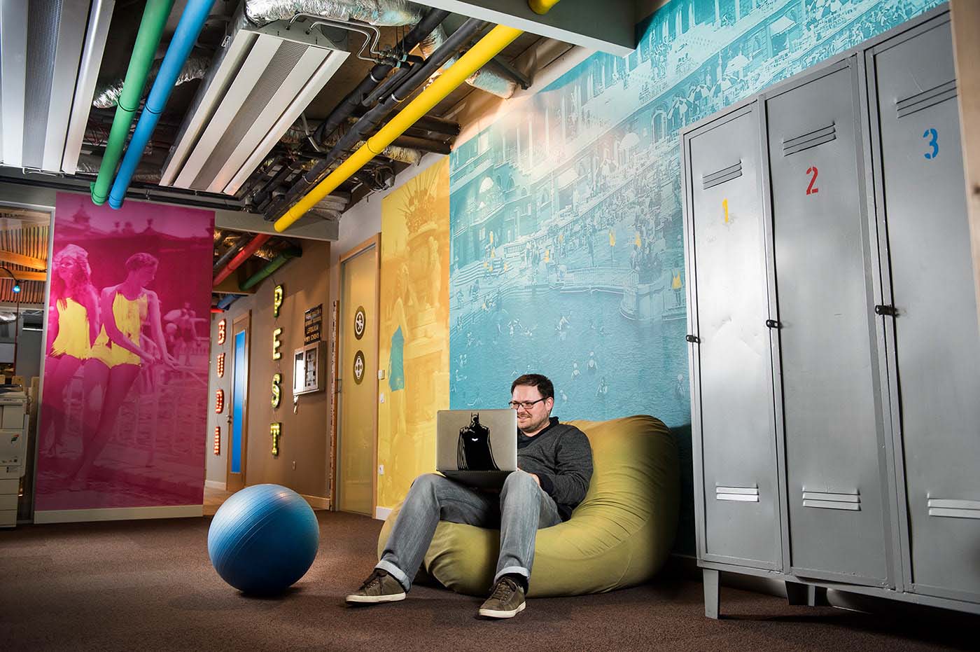 A Google Budapest iroda központi helyisége színes, nyomtatott faldekorációkkal, uszodai szekrényekkel, labdával és babzsákkal, amiben kényelmesen lehet dolgozni.