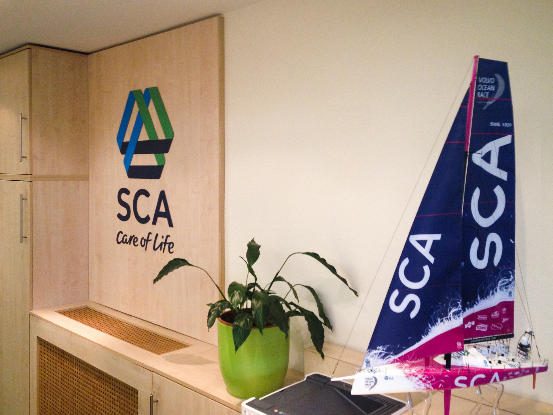 Az SCA irodája nagyméretű SCA logóval, szobanövénnyel és egy SCA arculati színeit viselő sportvitorlás makettjével az előtérben.