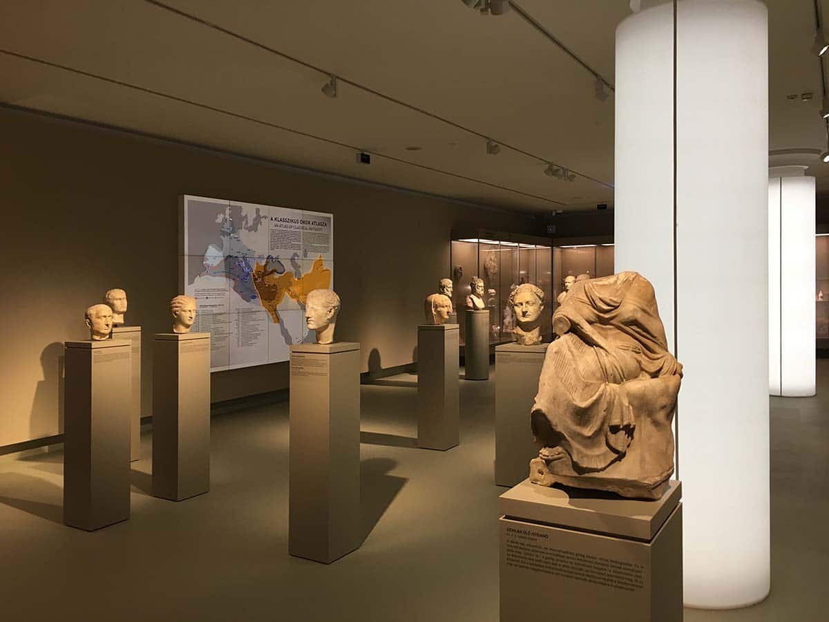 A Szépművészeti Múzeum ókor kiállítása korabeli antik szobrokkal az előtérben kellemes, fehér, világító oszlopokkal és látványos plexitérképpel a háttérben.