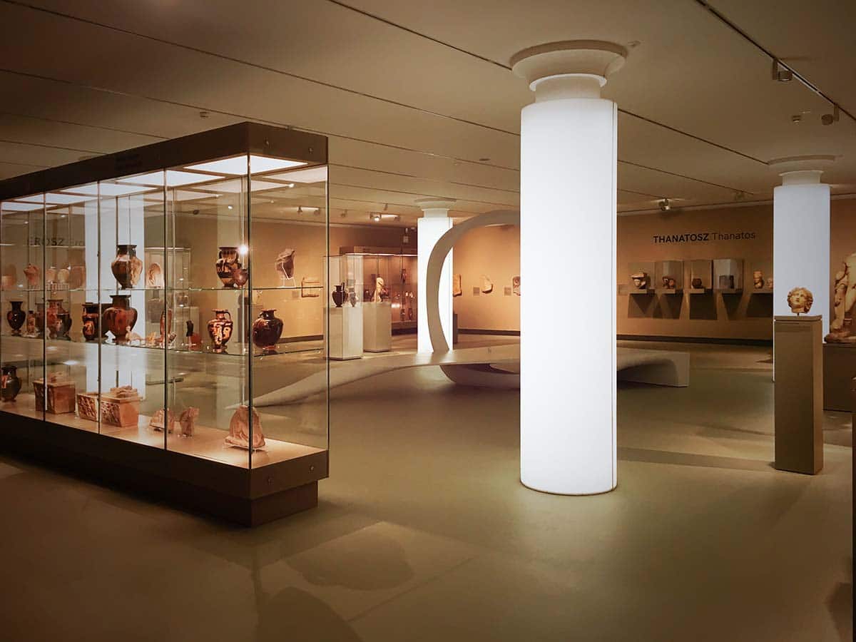 A Szépművészeti Múzeum ókor kiállítása. Antik vázák és edények, valamint más műtárgyak sorakoznak egy impozáns üvegvitrinben az előtérben, még kicsivel hátrébb, fehér, világító oszlopok egy organikus installáció, más vitrinek és szobrok látszanak.