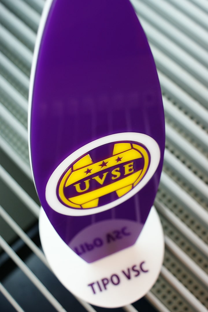 Egy frissen elkészült, visszafogott, mégis mutatós lila-fehér plexiplakett az UVSE sárga logójával a vágógép rácsain.