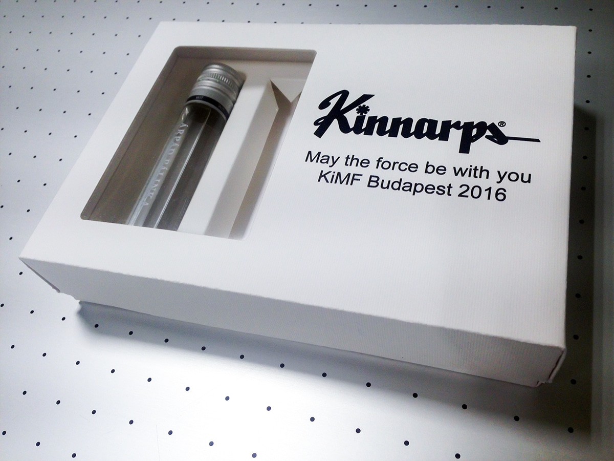 Fehér doboz a fekete Kinnarps logóval, a "May the force be with you KiMF Budapest 2016" felirattal és a kivágott ablakkal,a mi alatt különleges, fiolaalakú pálinkásüvegcse van.