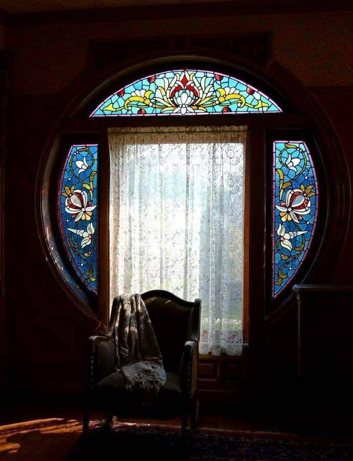 Kék alapon tekergőző virágmotívumos ólomüvegtáblák egy kör befoglaló formájú ablakkeretben. Átragyog rajtuk a színes fény a sötét szobába. Az ablak előtt kényelmes karosszék áll.